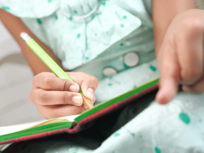 Für welche Rechtschreibthemen ist die Vokallänge wichtig - Mädchen mit grünem Klein schreibt mit grünem Stift in ein Buch mit rotem Einband.